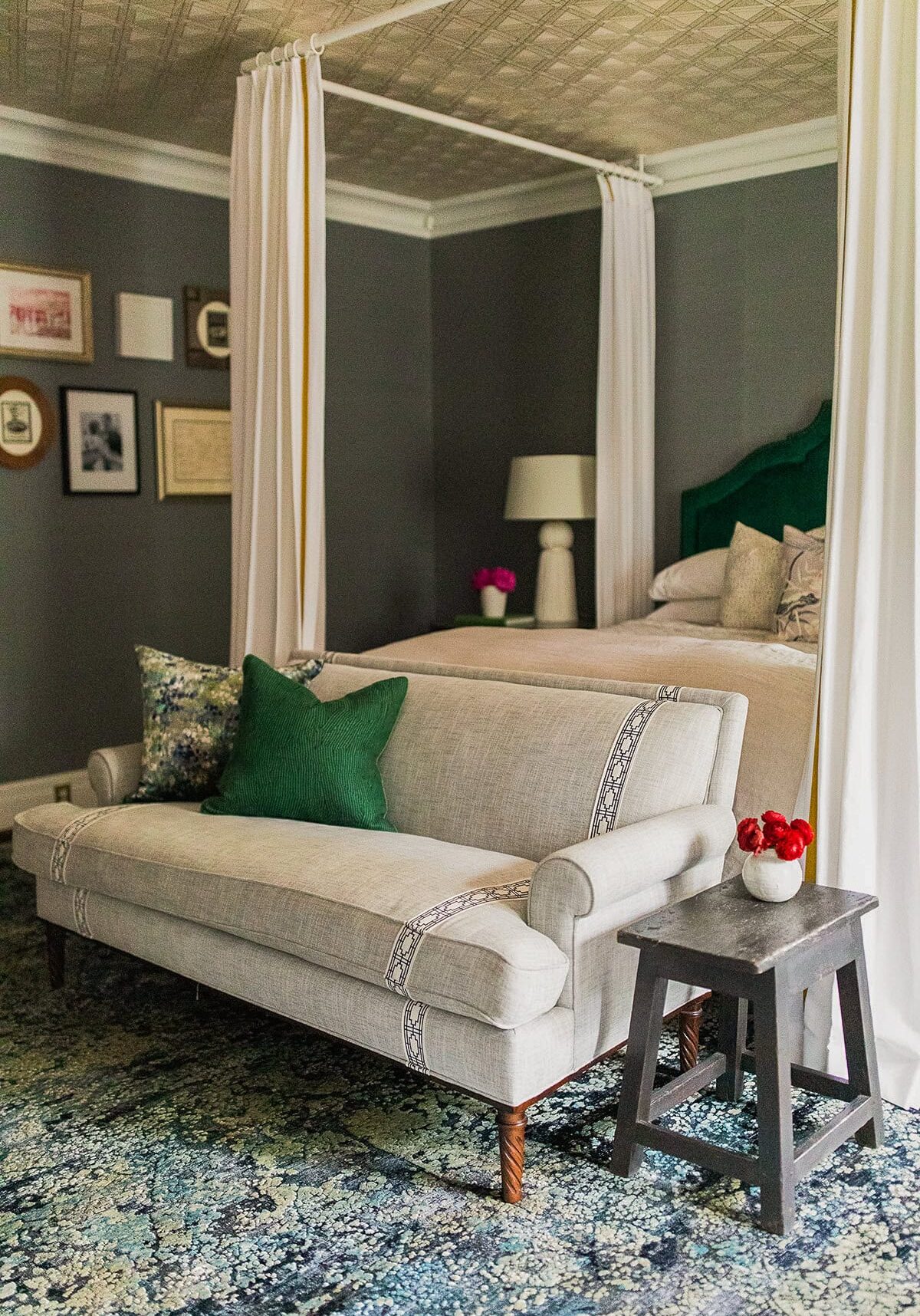 A gray upholstered settee with green and blue pillows sits at the foot of a canopy bed in a bedroom with dark walls, wall art, and a side table holding a vase of red flowers.