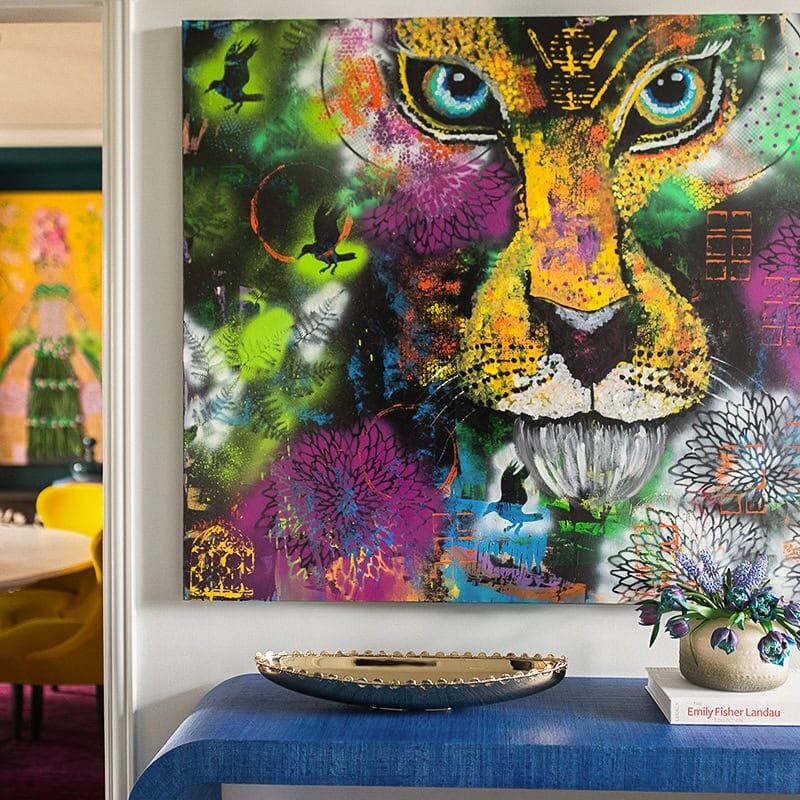 A vibrant, multi-colored graffiti-style painting of a lion’s face hangs above a blue textured console table with a gold bowl and purple tulips. In the background, a dining room with yellow chairs is visible through a doorway.