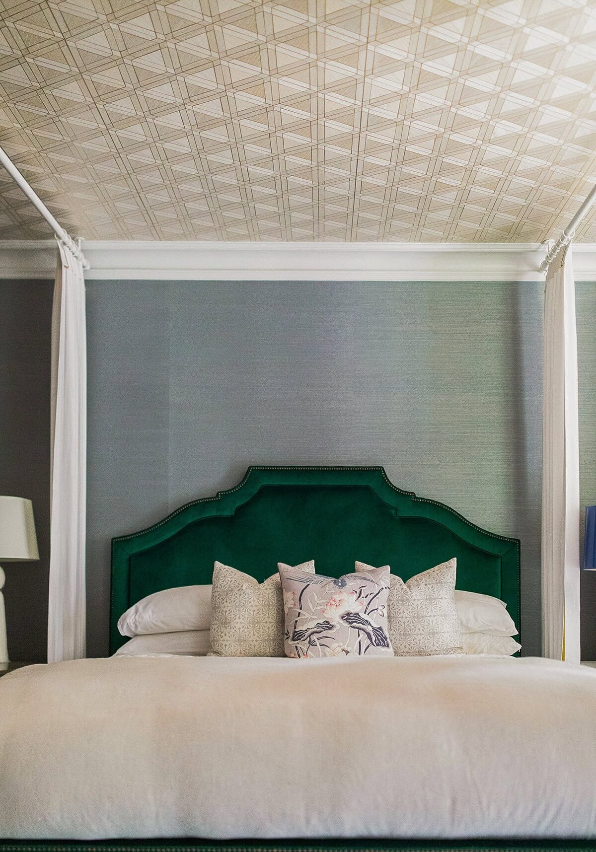 A neatly made bed with a green upholstered headboard, decorative pillows, white bedding, and a patterned ceiling above.