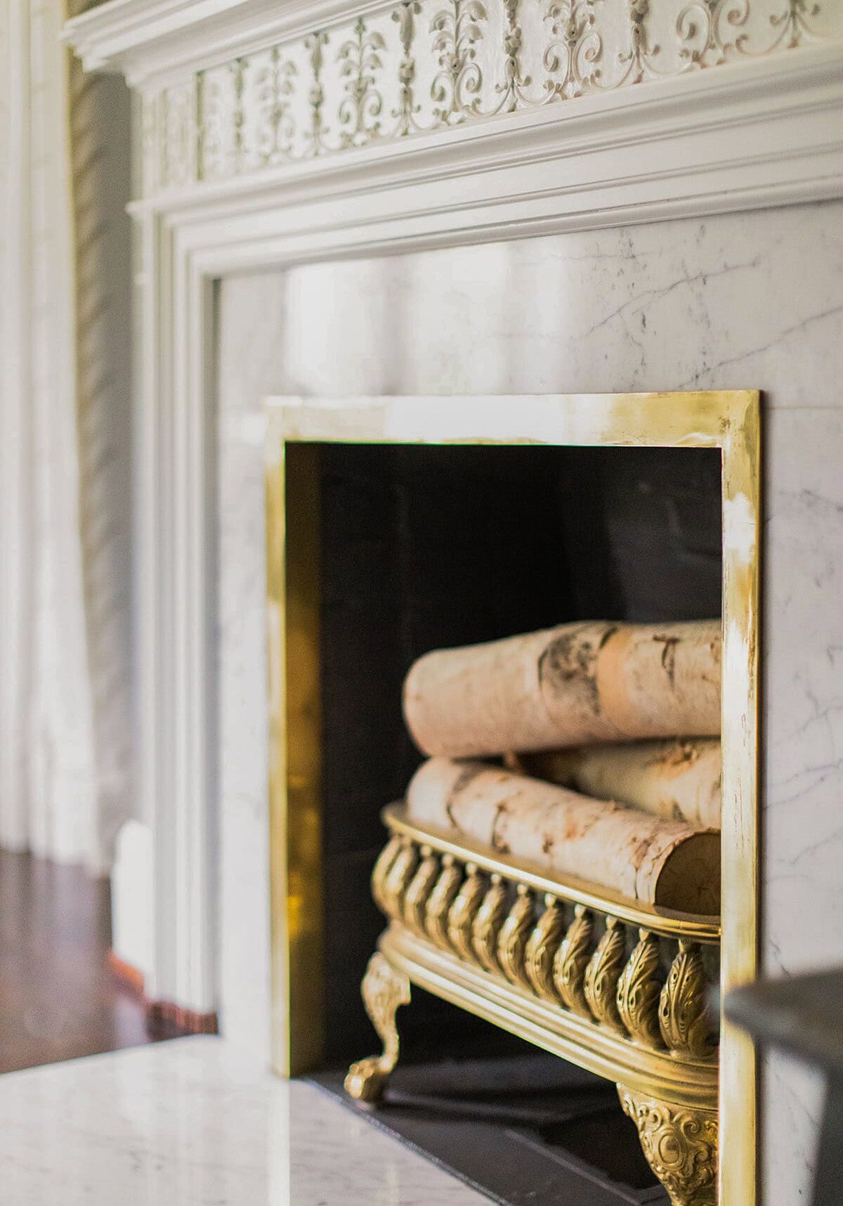 A marble fireplace with ornate trim, featuring a gold metal grate holding several large birch logs.