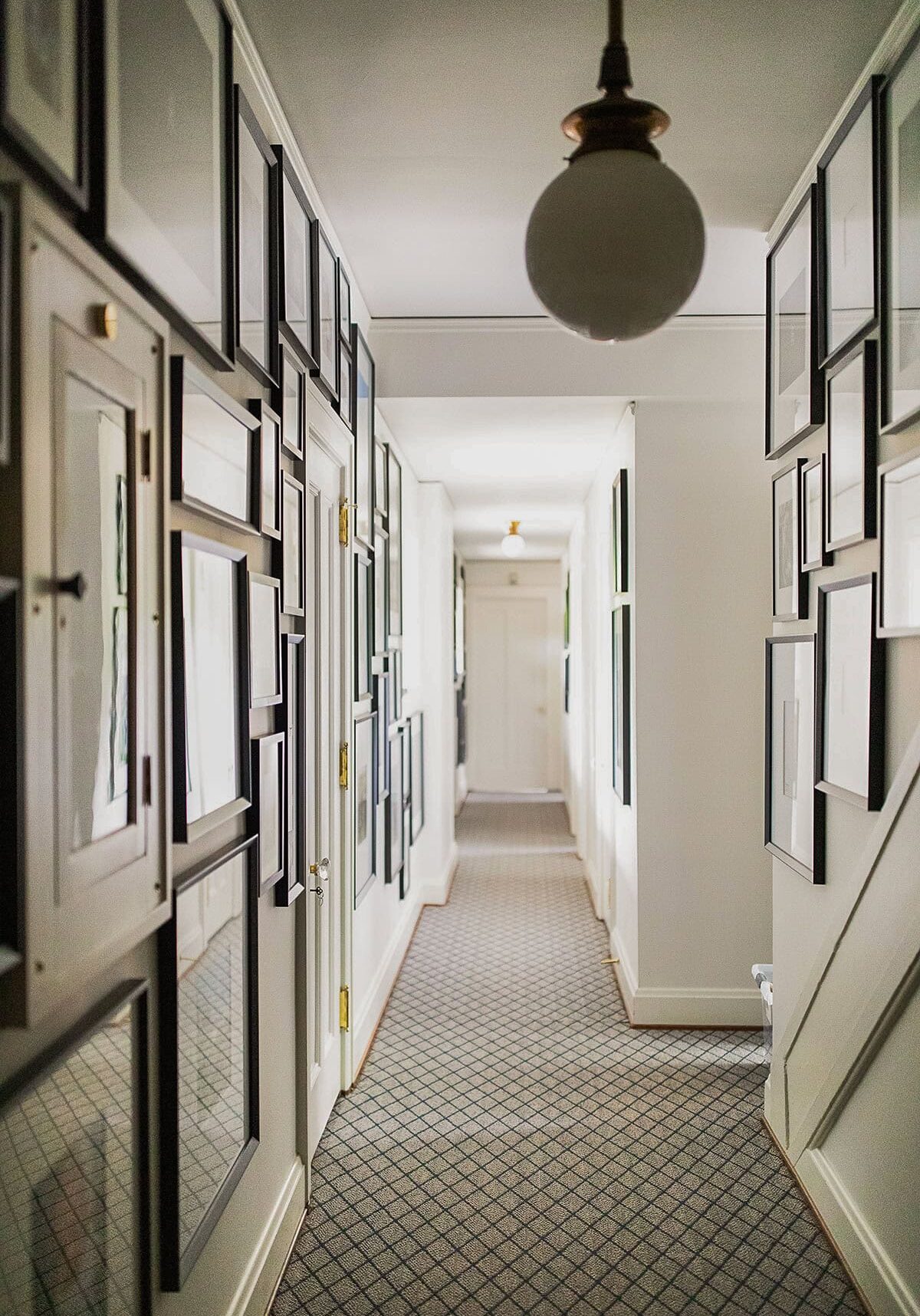 A narrow hallway with patterned carpet, white walls, and framed pictures hung on both sides, illuminated by a round ceiling light.