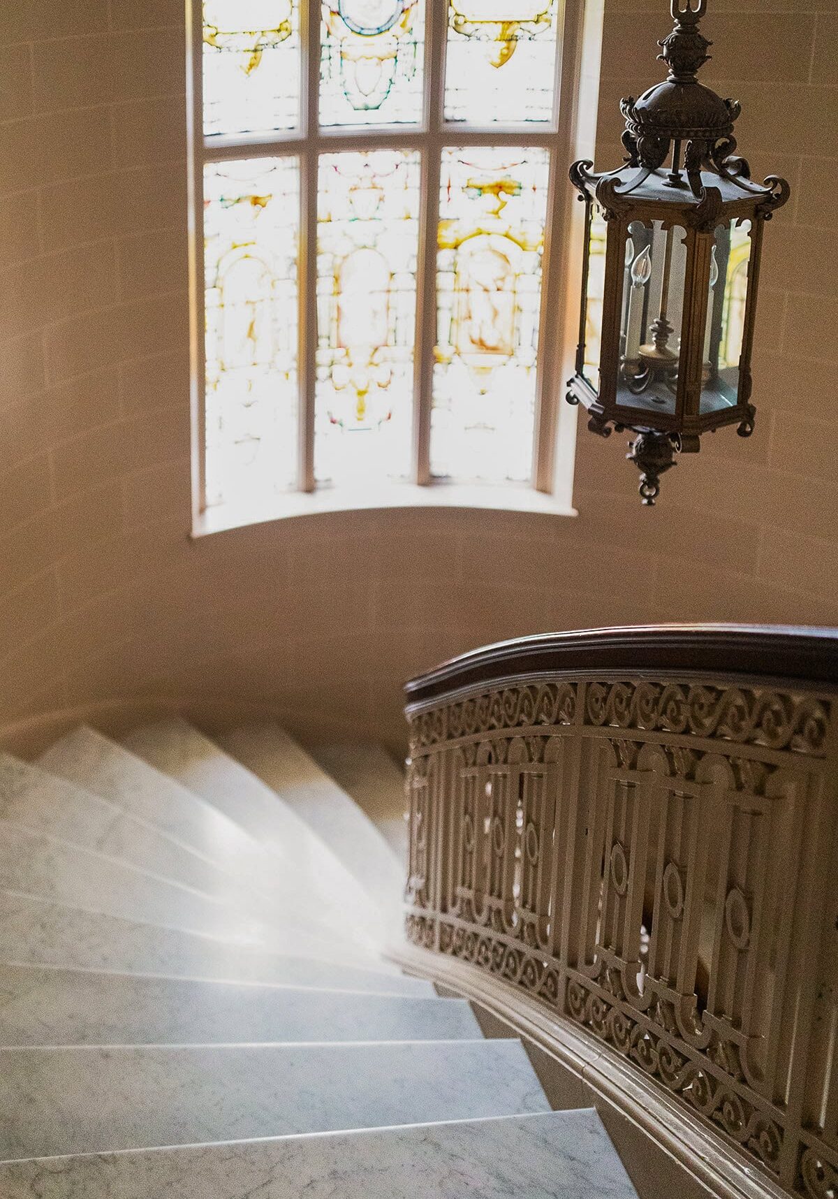 Curved marble staircase with ornate railing, large stained glass window, and hanging lantern overhead.