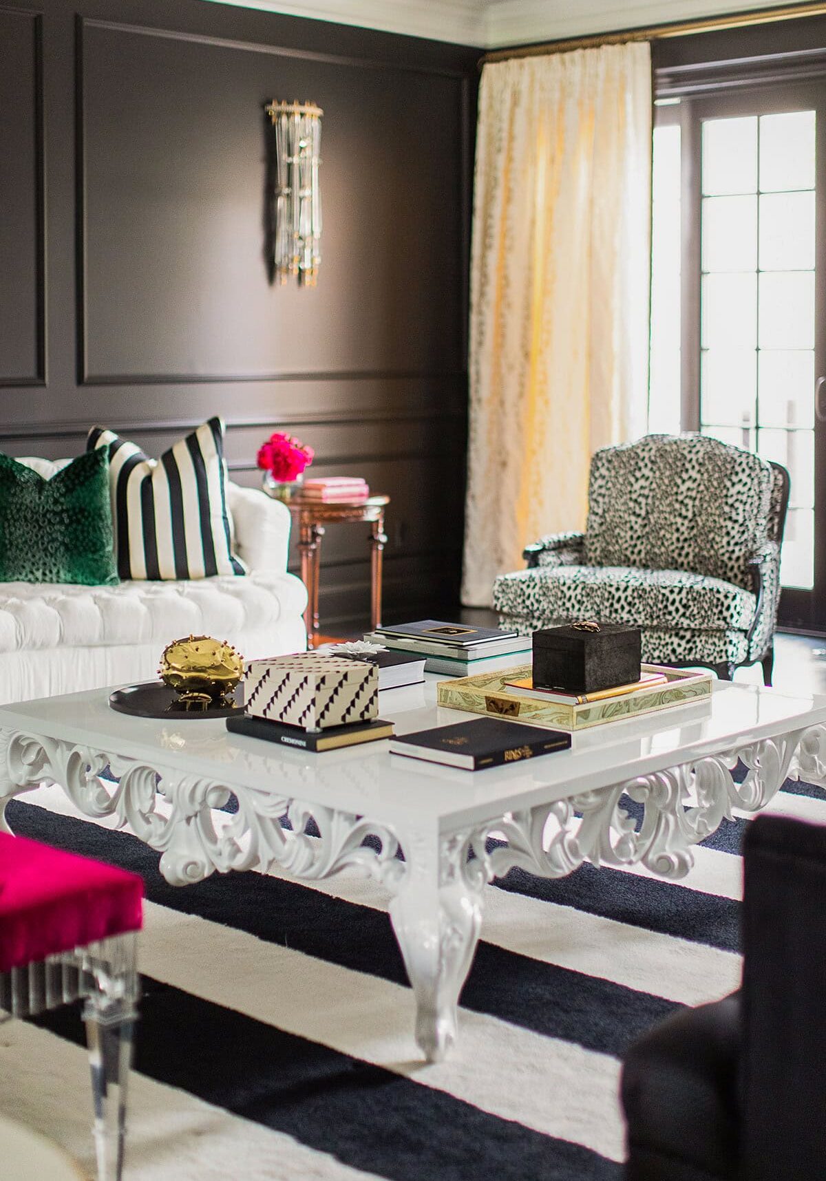 A living room with a white ornate coffee table, a white sofa with striped pillows, a patterned armchair, and black-and-white decor accents.