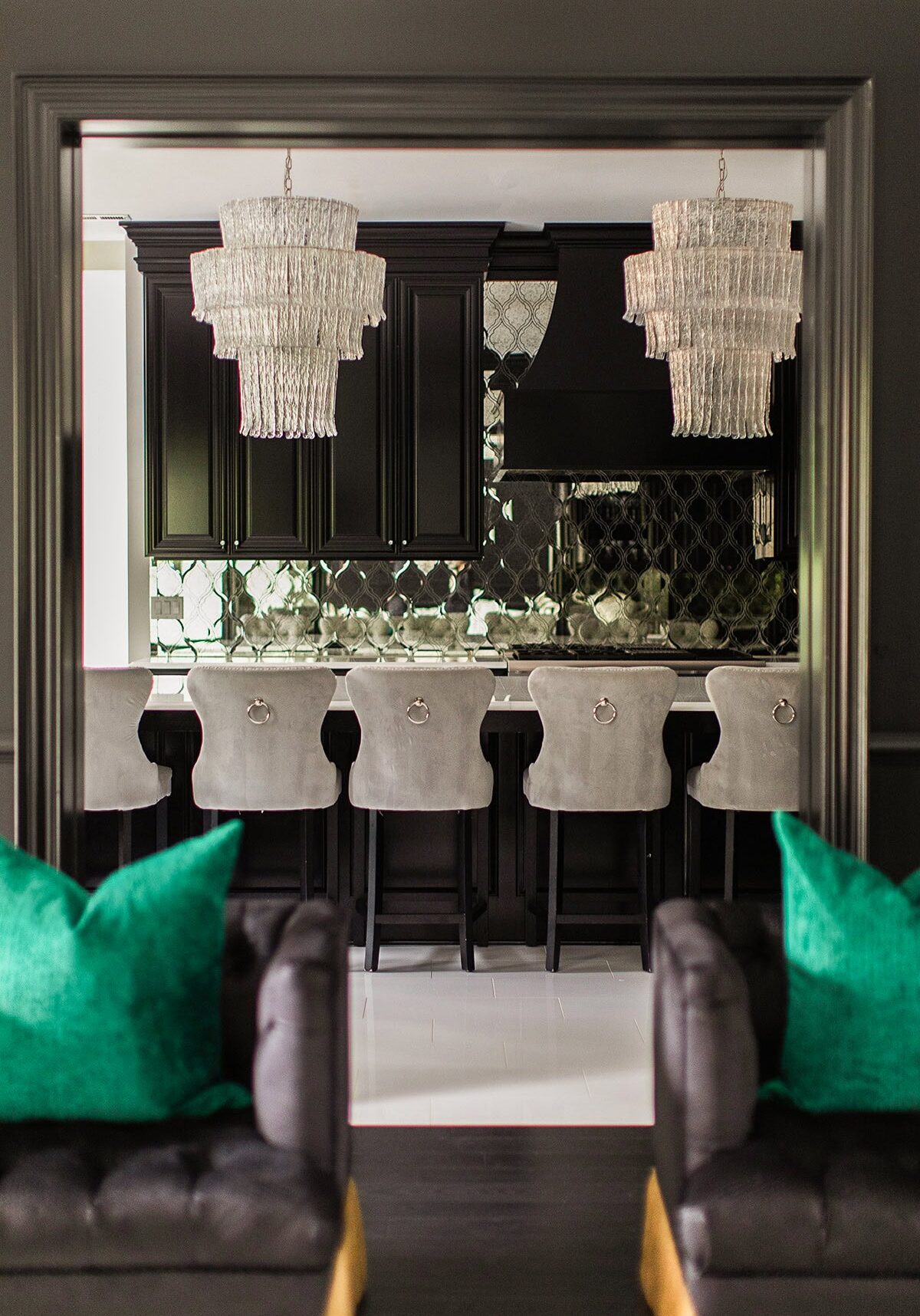 A modern kitchen features black cabinetry, mirrored backsplash, five gray barstools, two crystal chandeliers, and part of a living area with dark sofas and green pillows in the foreground.