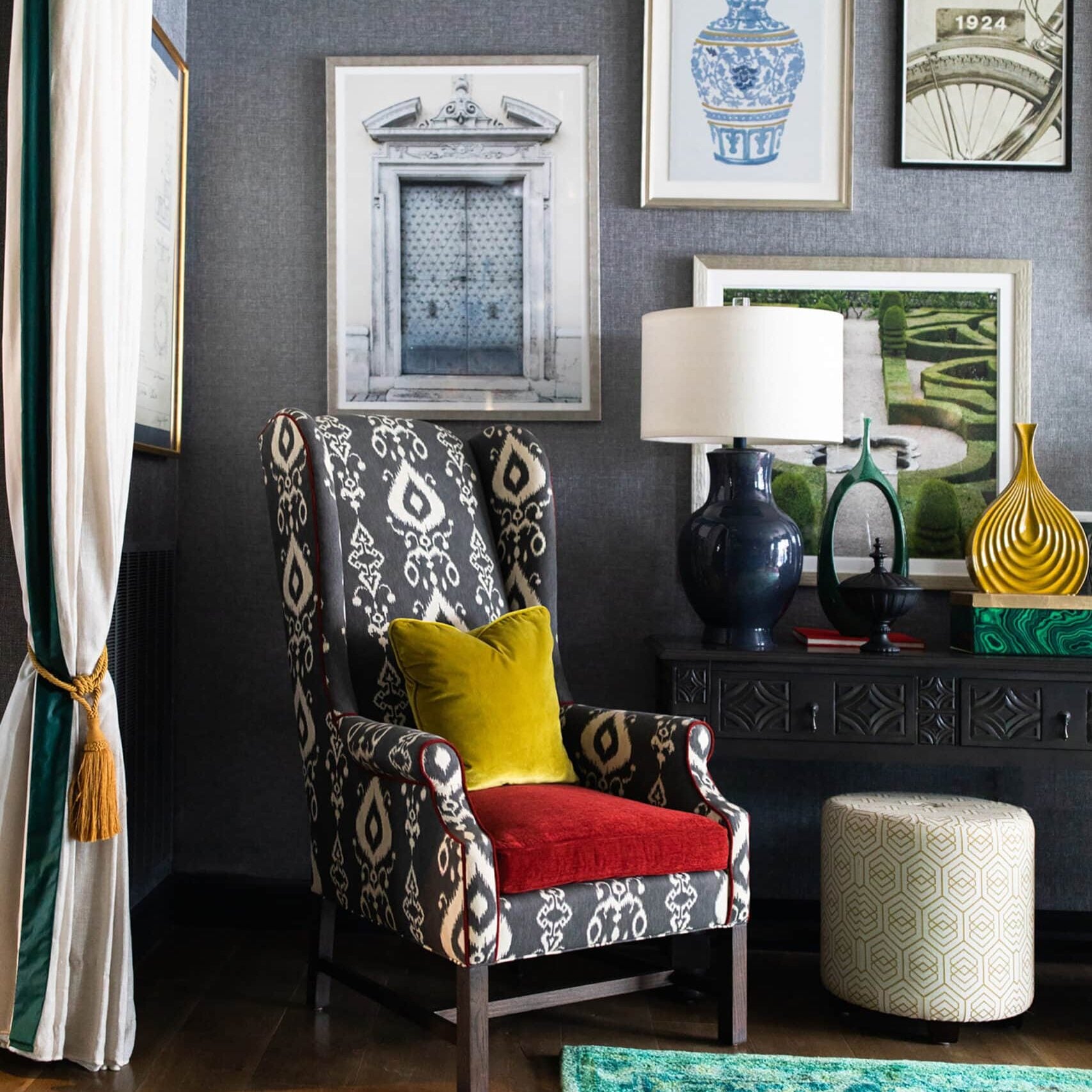 A patterned armchair with a yellow pillow sits beside a modern table, lamp, and decor; framed art hangs on a gray wall above a green rug and pouf.