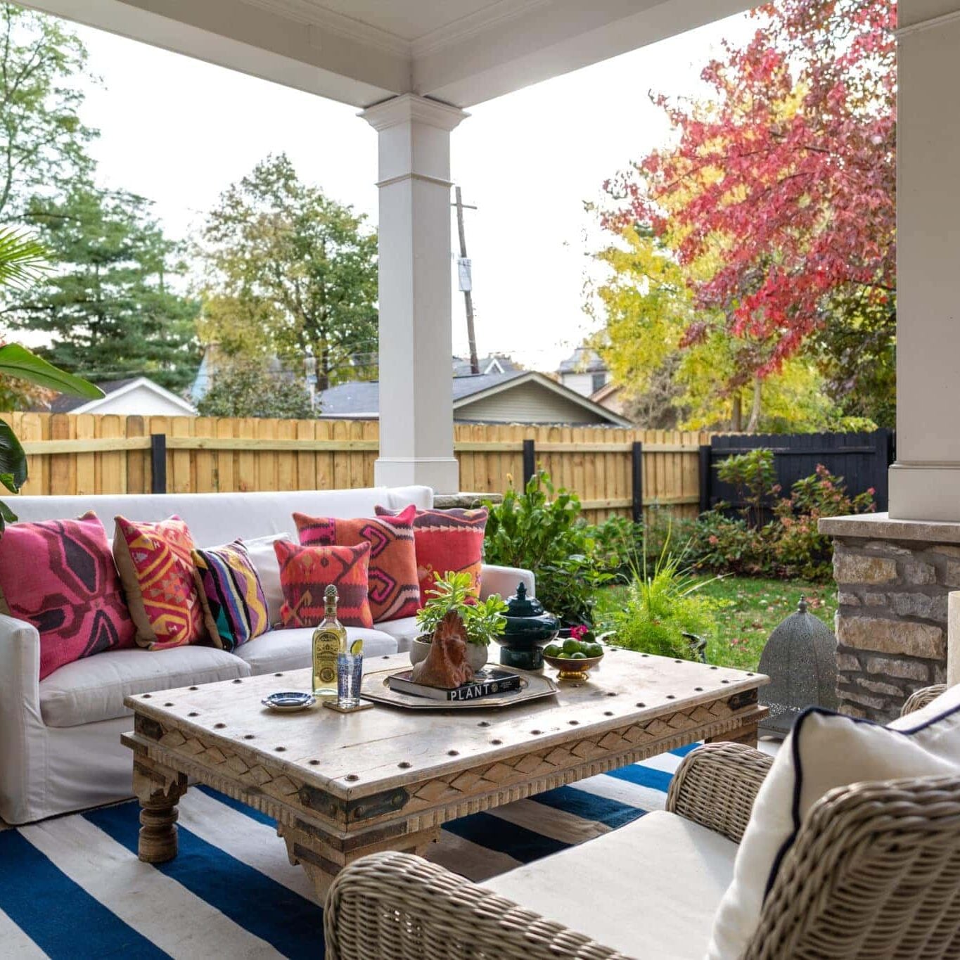 A covered patio designed by Crimson Design Group features a white sofa with colorful cushions, wicker chairs, and a wooden coffee table, all set beside a fenced backyard adorned with autumn-colored trees.