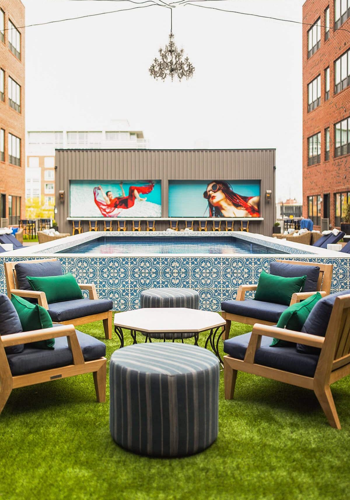 Outdoor lounge area with cushioned chairs and ottomans on artificial grass, facing a tiled pool and digital screens displaying images of women swimming.