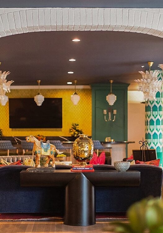 A modern lounge with eclectic decor, featuring a black sofa, ceramic vases, decorative objects, and green chairs under ornate light fixtures and patterned walls.