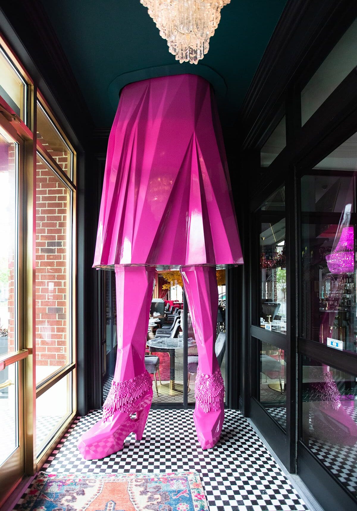 Large pink sculpture of legs in high-heeled shoes and a skirt stands under a chandelier on a black-and-white checkered floor near a window and mirrored wall.