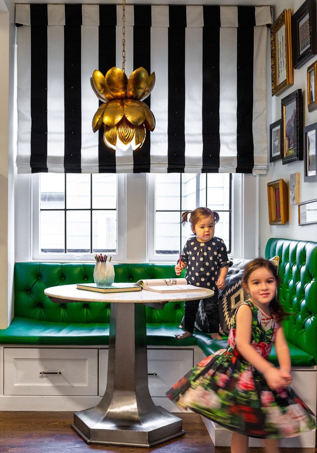 Two young girls play in a brightly lit room with a green tufted bench, round table, striped window shade, and a gold flower-shaped pendant light overhead.