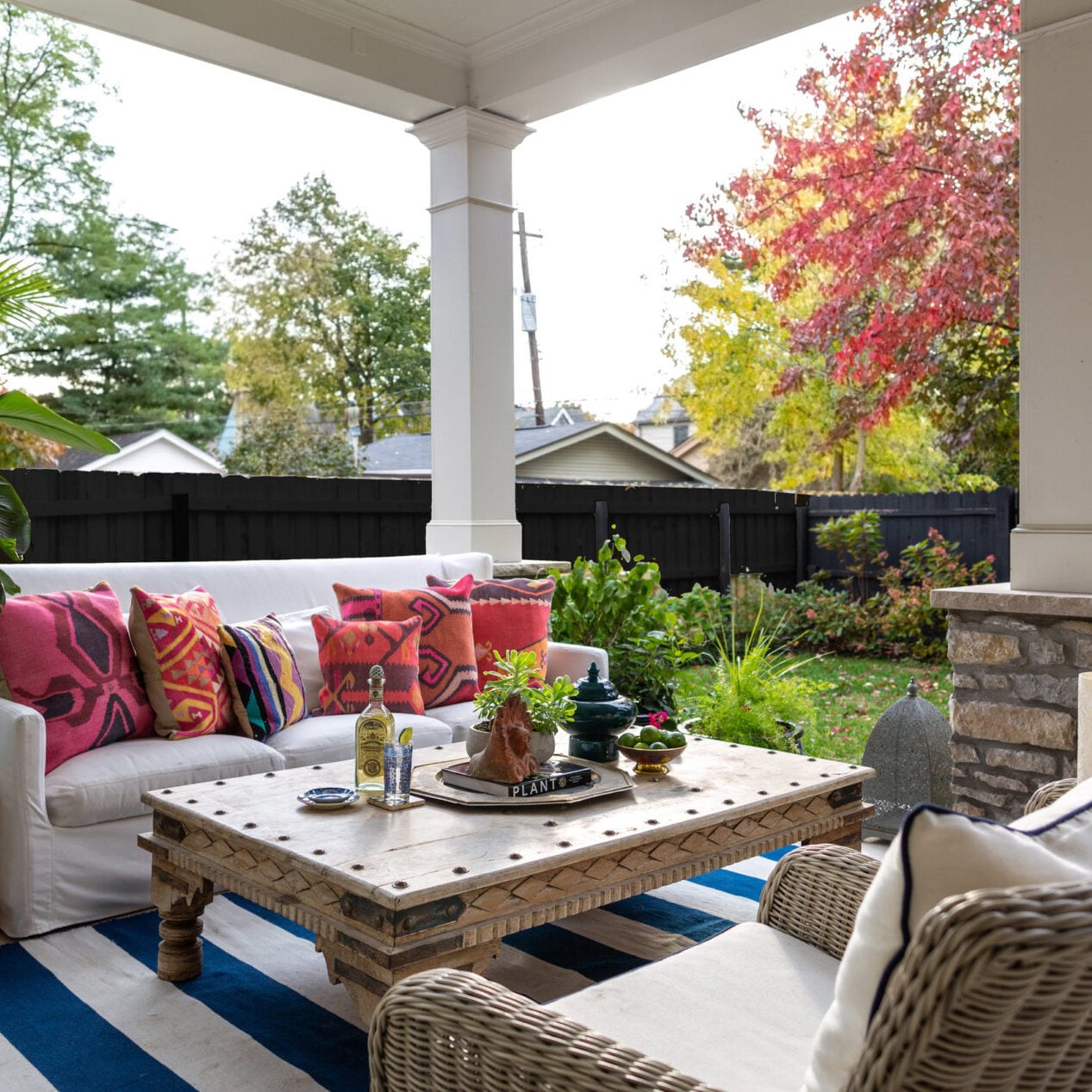 A covered patio designed by Crimson Design Group features a white sofa, colorful pillows, a wooden coffee table, wicker chairs, a striped rug, and a stunning garden view with autumn trees.