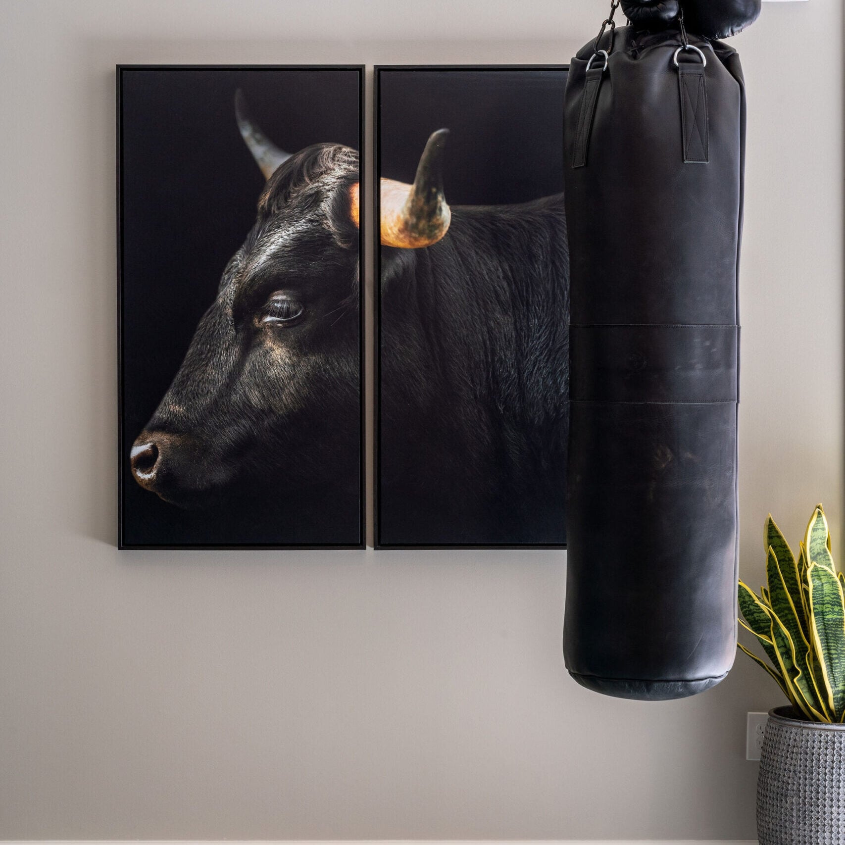 A black punching bag with boxing gloves hangs in front of a wall with a two-panel bull portrait; a plant sits on the floor nearby.