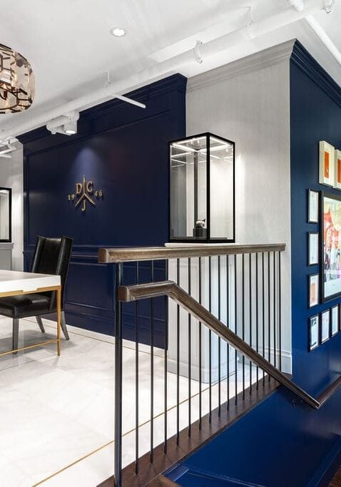 Modern office entryway with navy blue accent walls, a white desk with a black chair, wall art, pendant lights, and a staircase with a sleek railing.