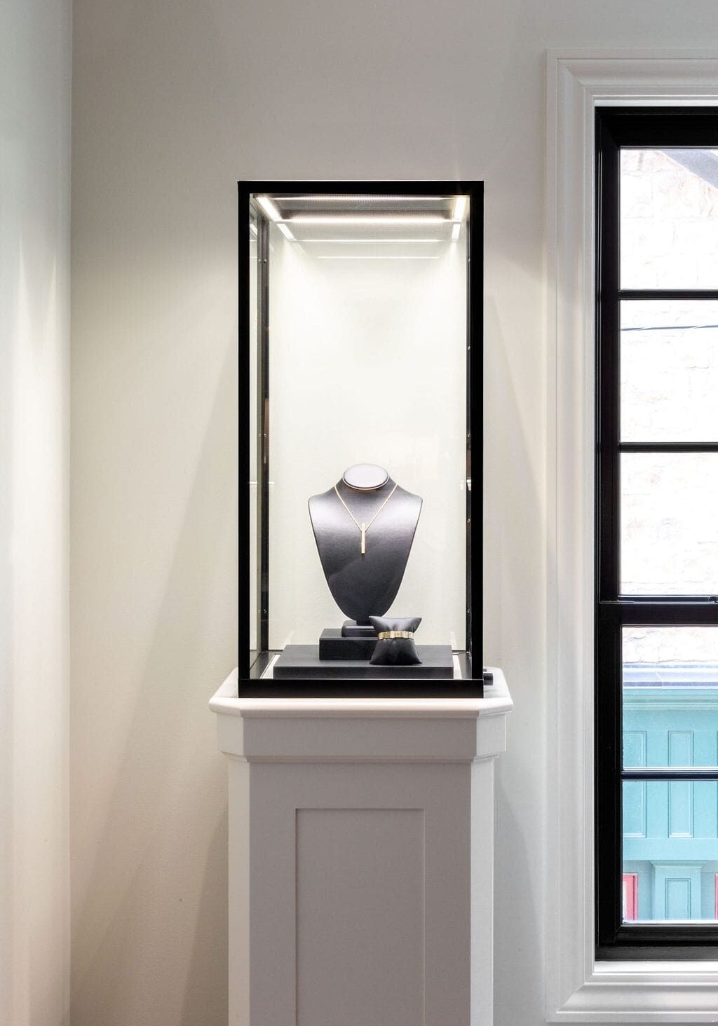 A jewelry display case holds a necklace and a bracelet on stands, set against a white wall next to a window with black trim.