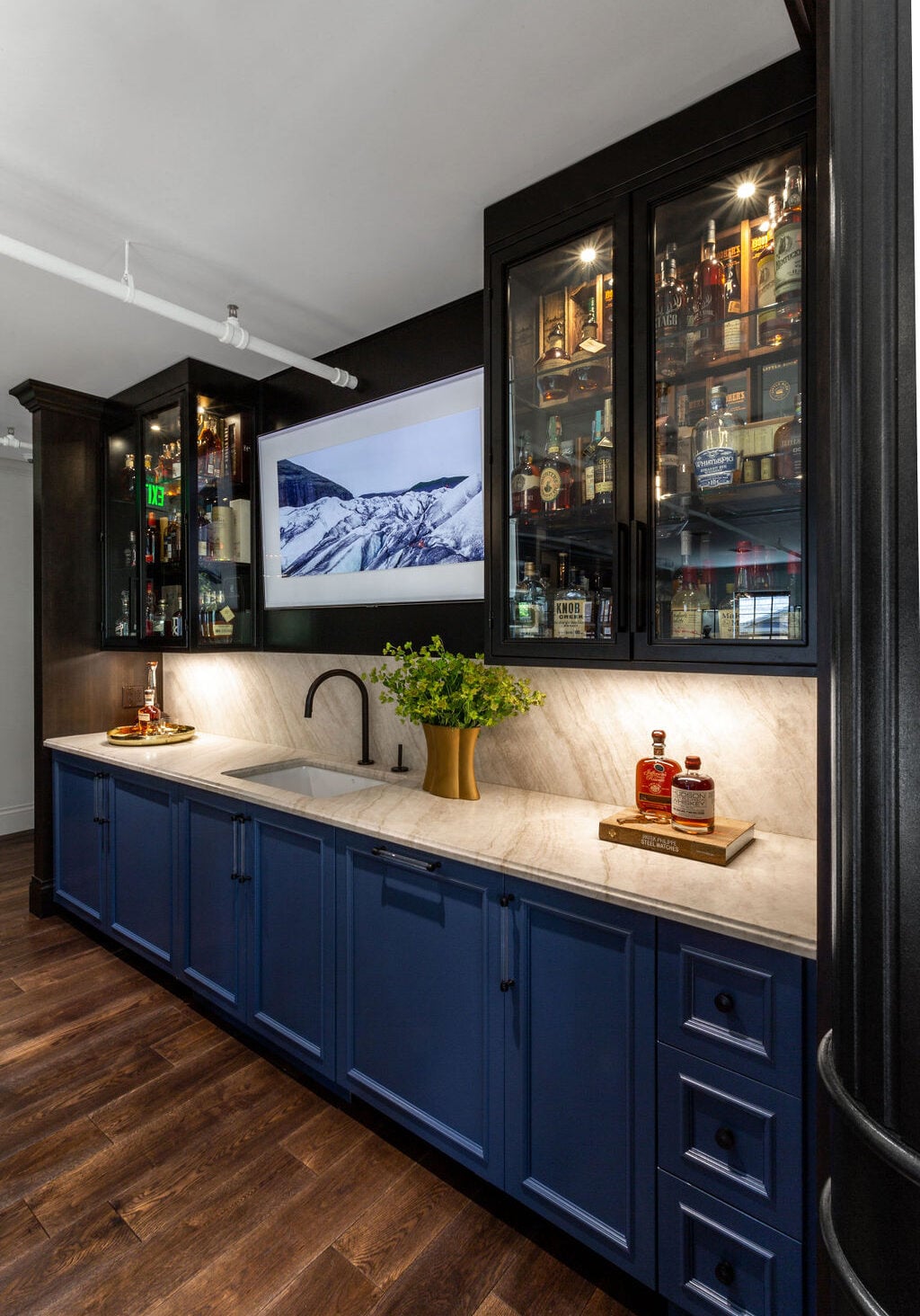 A modern home bar with blue cabinets, a marble countertop, glass-fronted upper cabinets filled with bottles, a sink, and a wall-mounted TV above the counter.