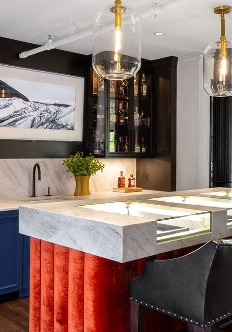 Modern home bar with marble countertop, red velvet bar front, black leather bar stools, glass cabinets with liquor bottles, and a wall-mounted landscape photo above the sink.