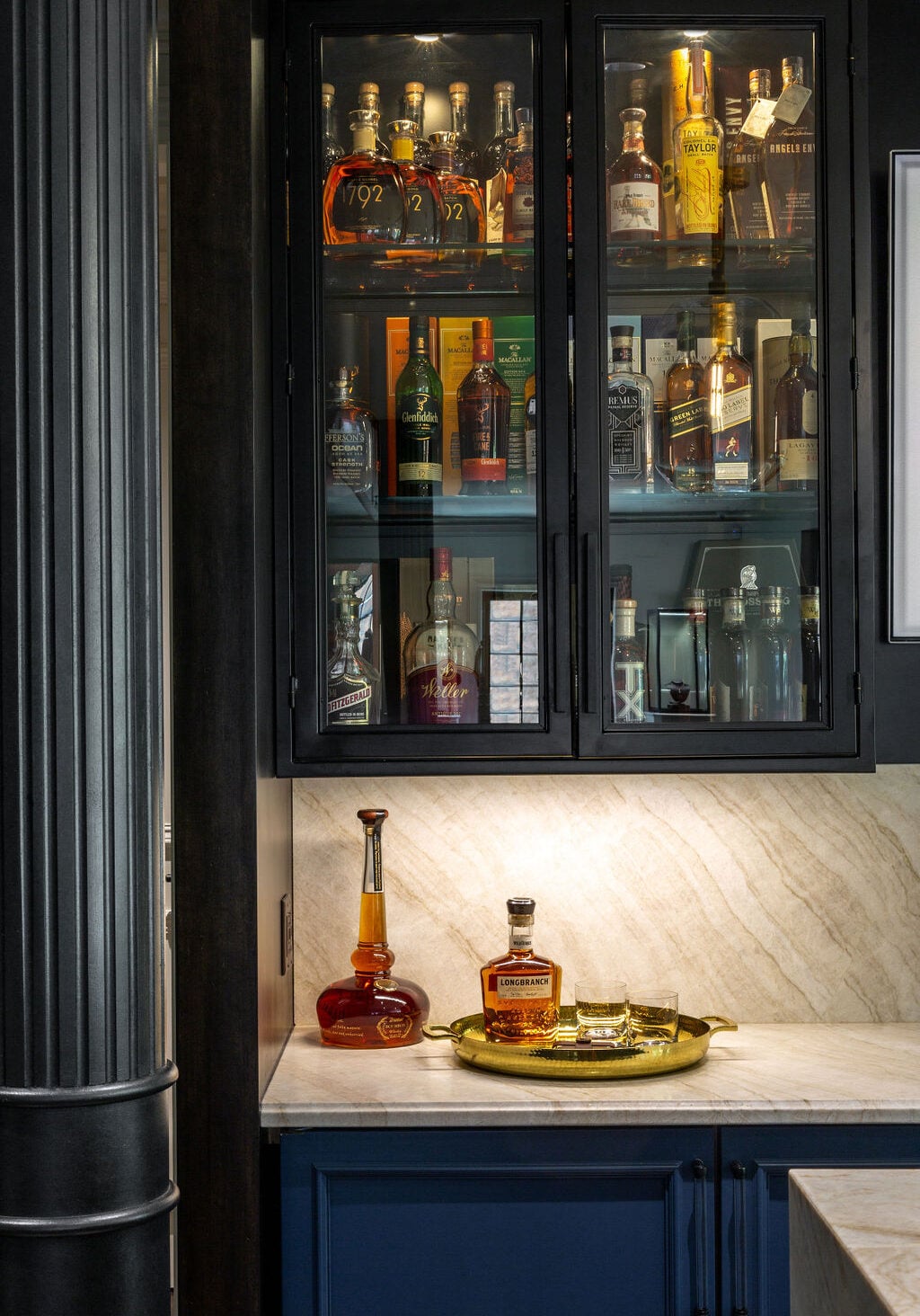 Glass cabinet with various liquor bottles displayed above a marble countertop, which holds a decanter, a bottle of whiskey, and two glasses on a gold tray.