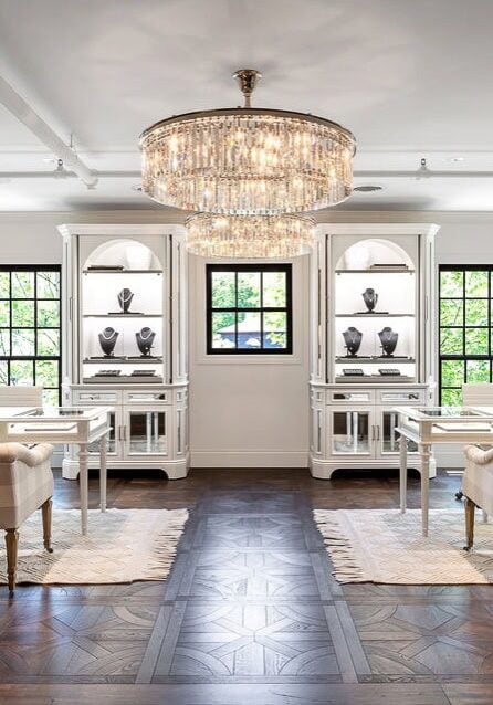 A bright, elegant showroom with two plaid sofas, white tables, display cabinets with jewelry, a large chandelier, and framed art on the walls.