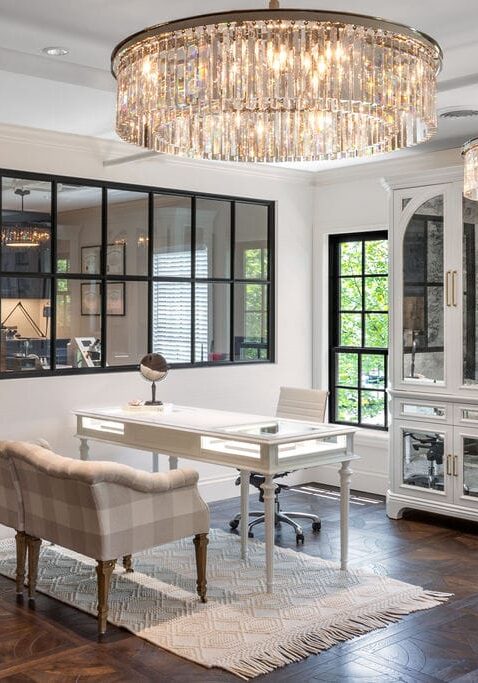 A modern home office with a white desk, a plaid upholstered bench, a white chair, glass-front cabinets, wood floors, and two large crystal chandeliers.