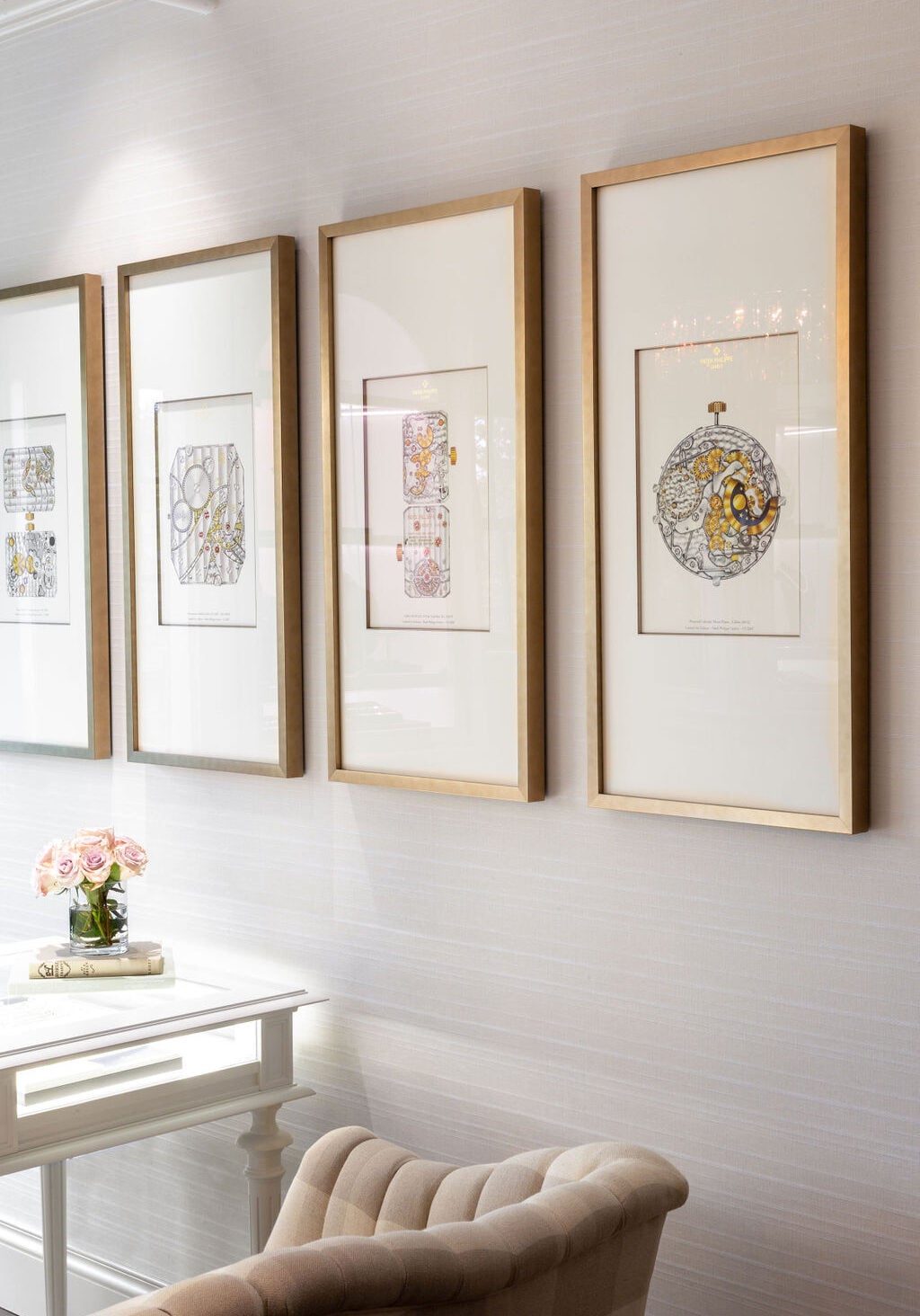 Four framed abstract art prints hang on a light-colored wall above a white desk with a vase of pink flowers and a beige upholstered chair in front.