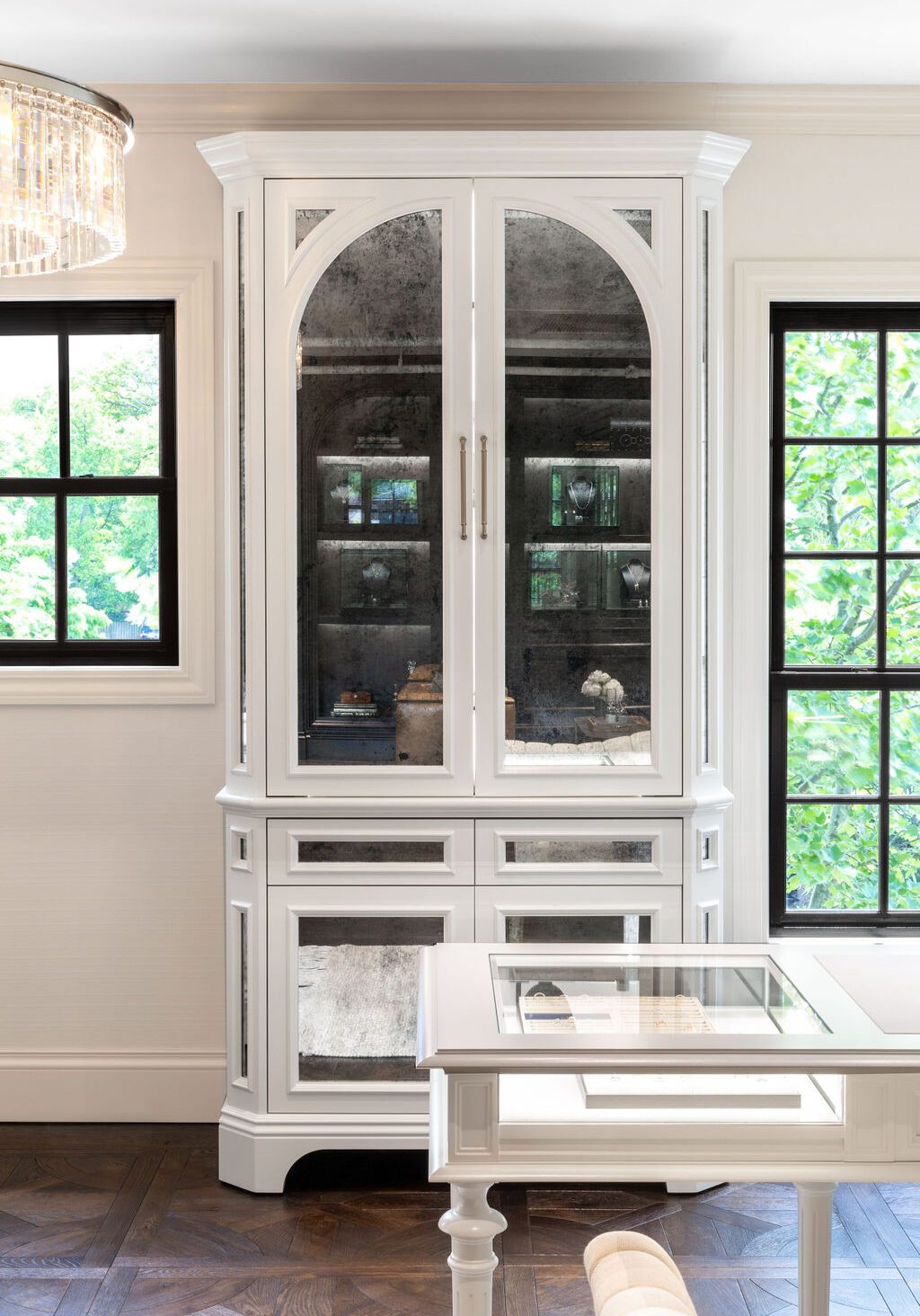 A white display cabinet with mirrored doors stands between two black-framed windows in a bright room with dark wood flooring.