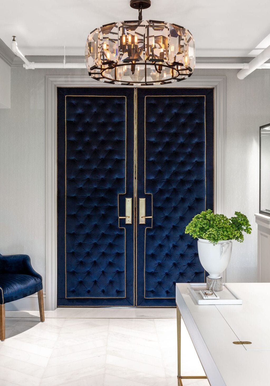 Double doors upholstered in deep blue tufted fabric with gold accents, beneath a modern chandelier. A white table with a potted plant sits in the foreground.