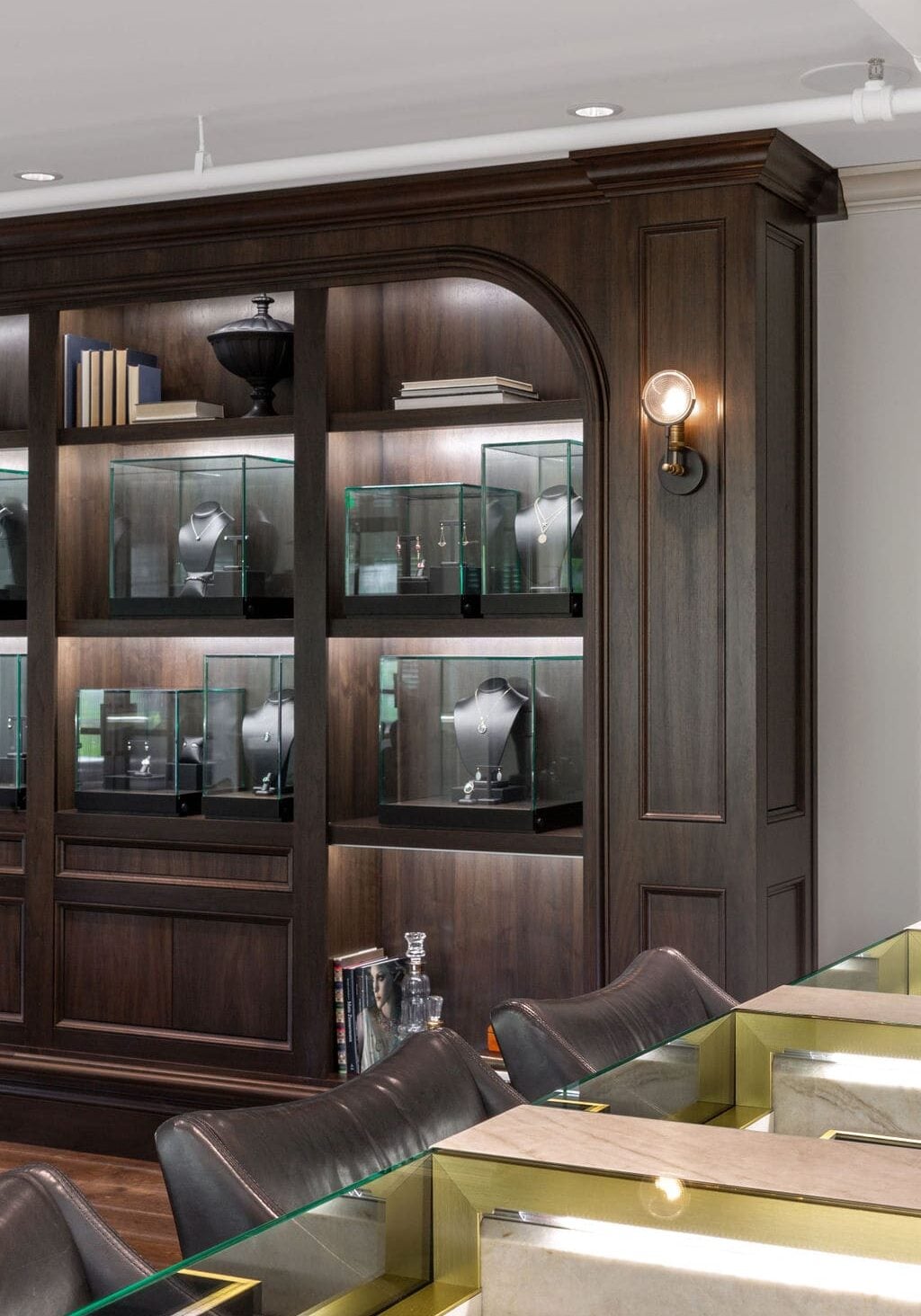 A wooden display wall with glass cases showcasing jewelry, books on the top shelf, and black chairs arranged by a glass-topped counter.