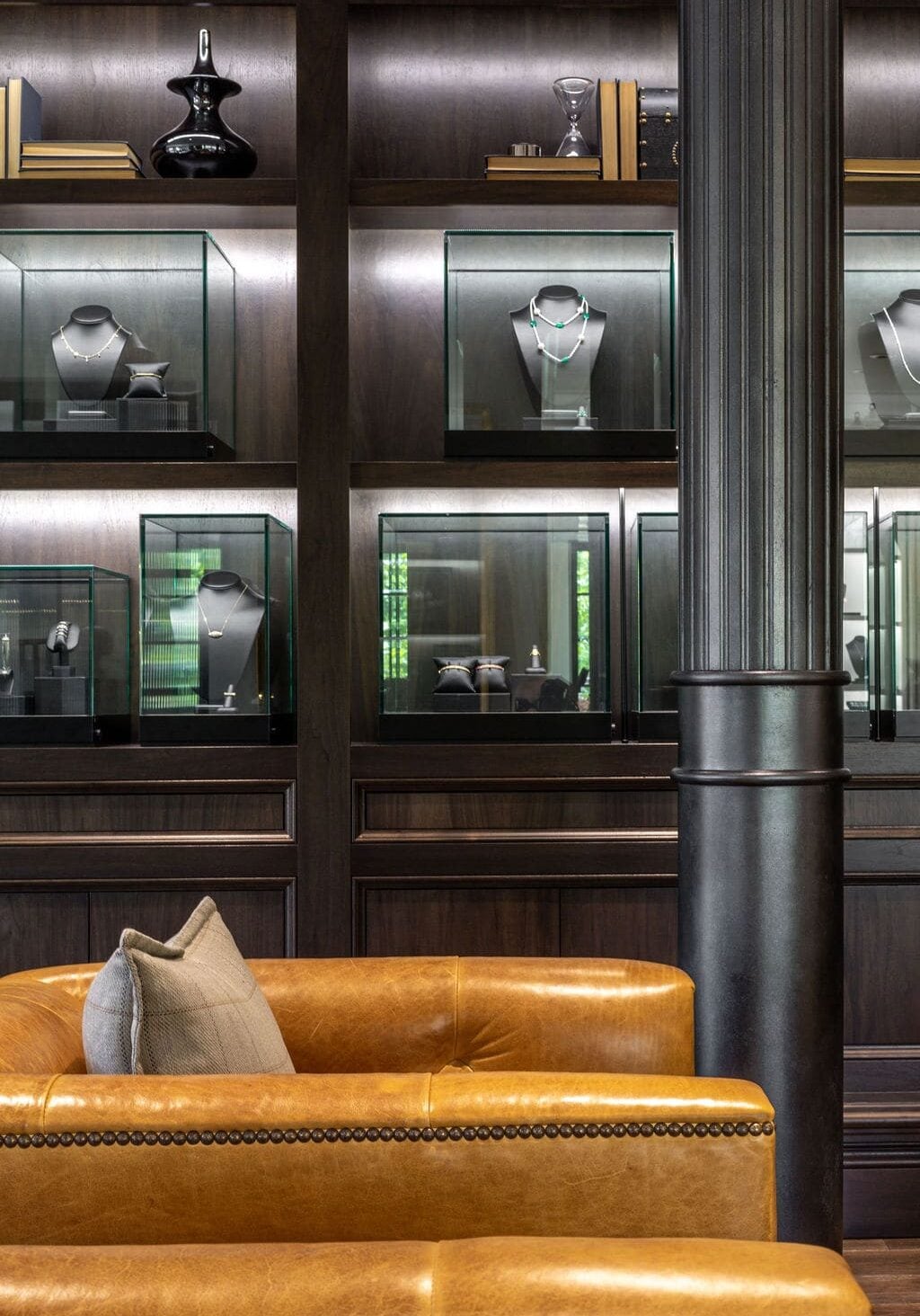 Glass display cases with jewelry on shelves are set into a dark wood wall behind a tan leather sofa and a black column.