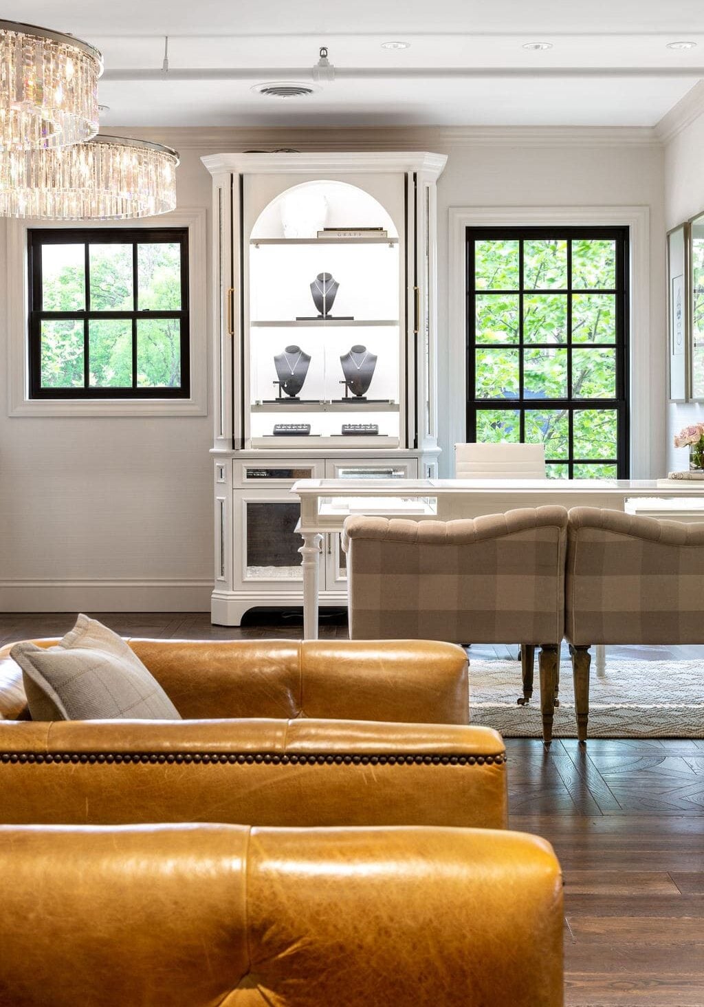 A living room features tan leather chairs, a plaid upholstered bench, a white display cabinet with jewelry busts, and large black-trim windows with greenery outside.