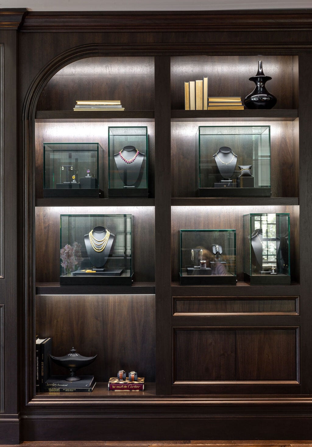 Dark wooden display shelves featuring glass cases with jewelry, books, and decorative items, all illuminated by built-in lighting.