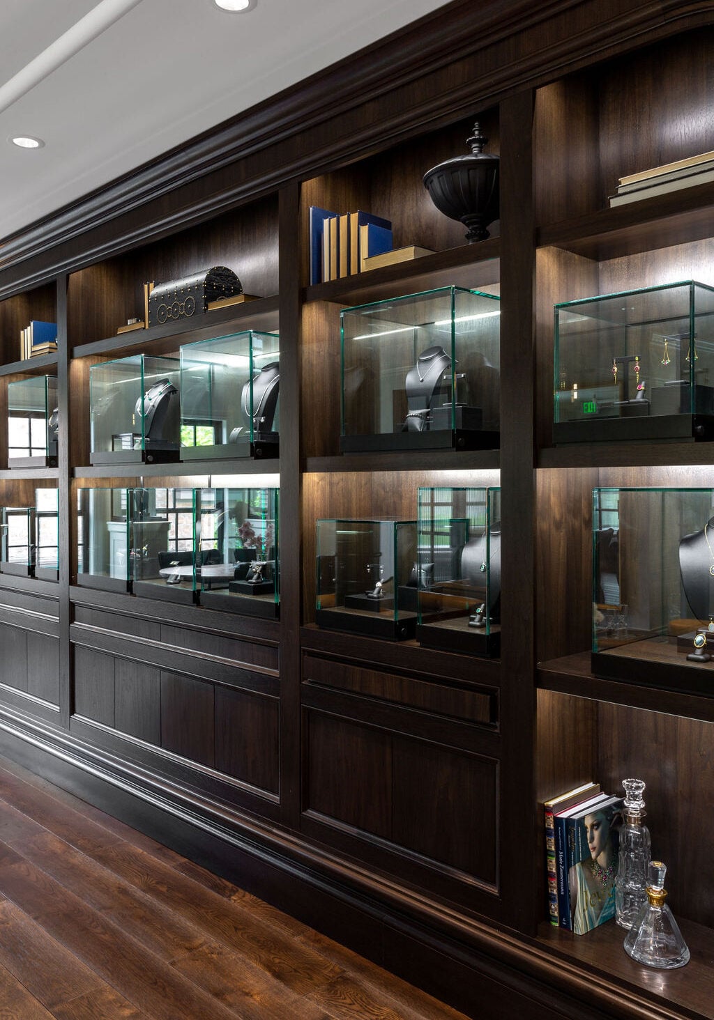 A dark wood display wall features glass cases with jewelry on stands, books, and decorative objects in a well-lit room with hardwood floors.