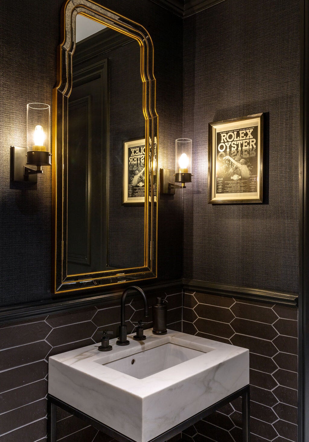 Modern bathroom with dark textured walls, rectangular marble sink, gold-framed mirror, wall sconces, and framed "Rolex Oyster" advertisement on the wall.