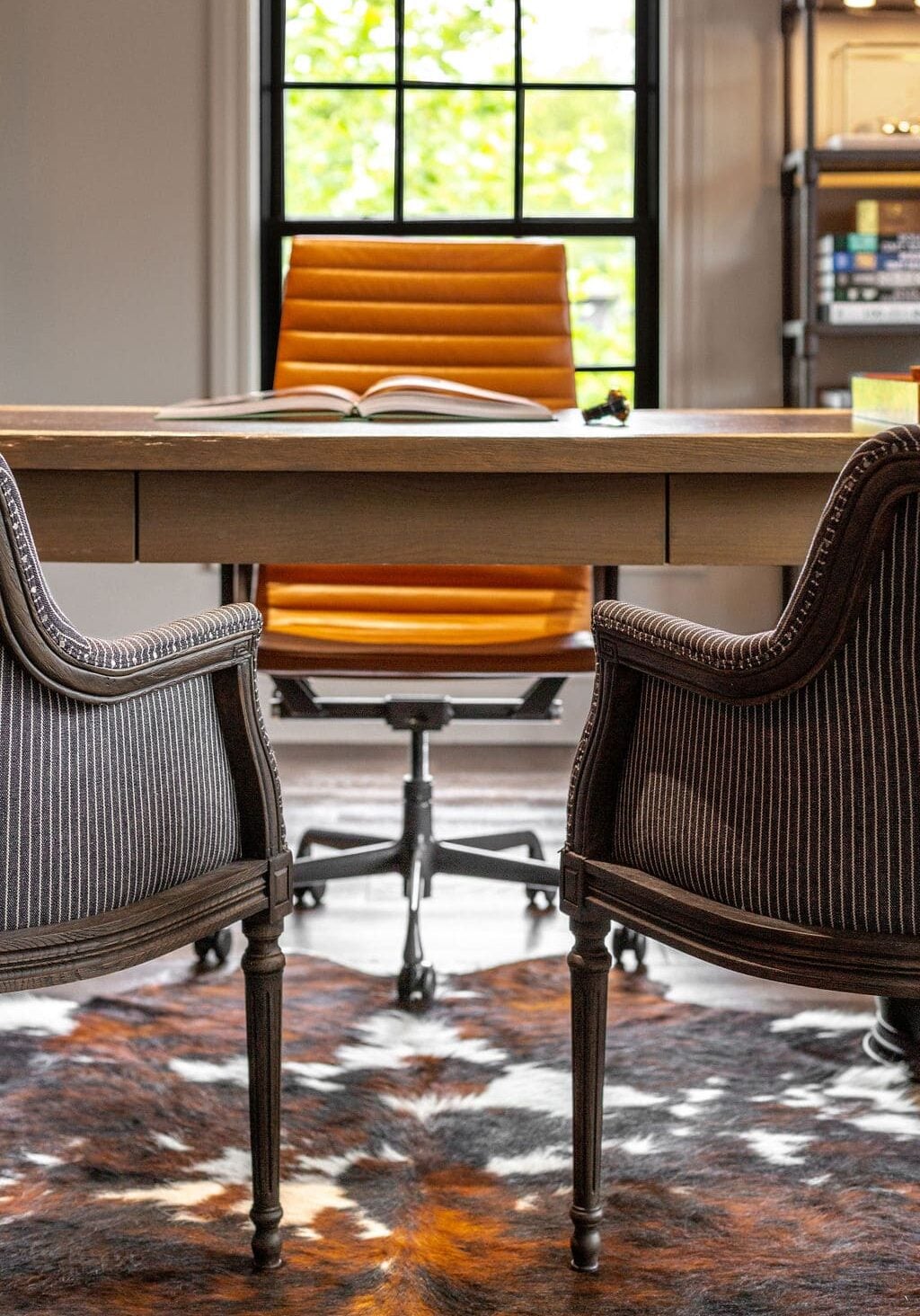 A modern office with an orange desk chair, two striped armchairs, a wooden desk with an open book, and a large window in the background.