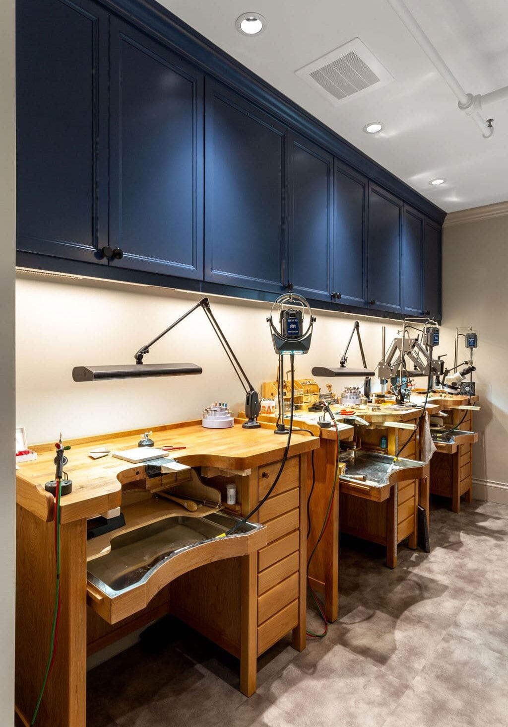 A row of wooden jeweler's workbenches with tools and equipment under blue cabinets and bright task lighting.