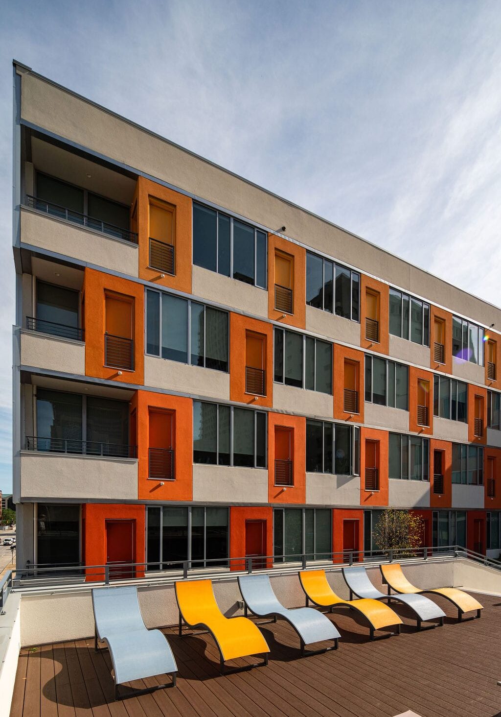 A modern apartment building with orange and yellow window shutters, seen behind a rooftop deck with yellow and gray lounge chairs.
