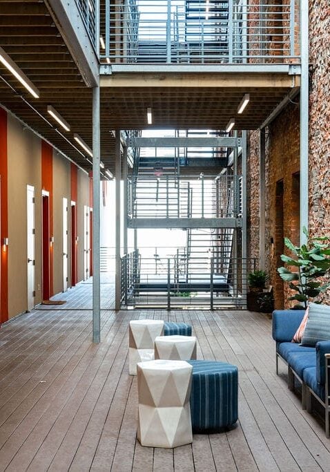 A modern indoor corridor with red doors, exposed brick wall, metal stairs, wood flooring, potted plants, and contemporary seating area.