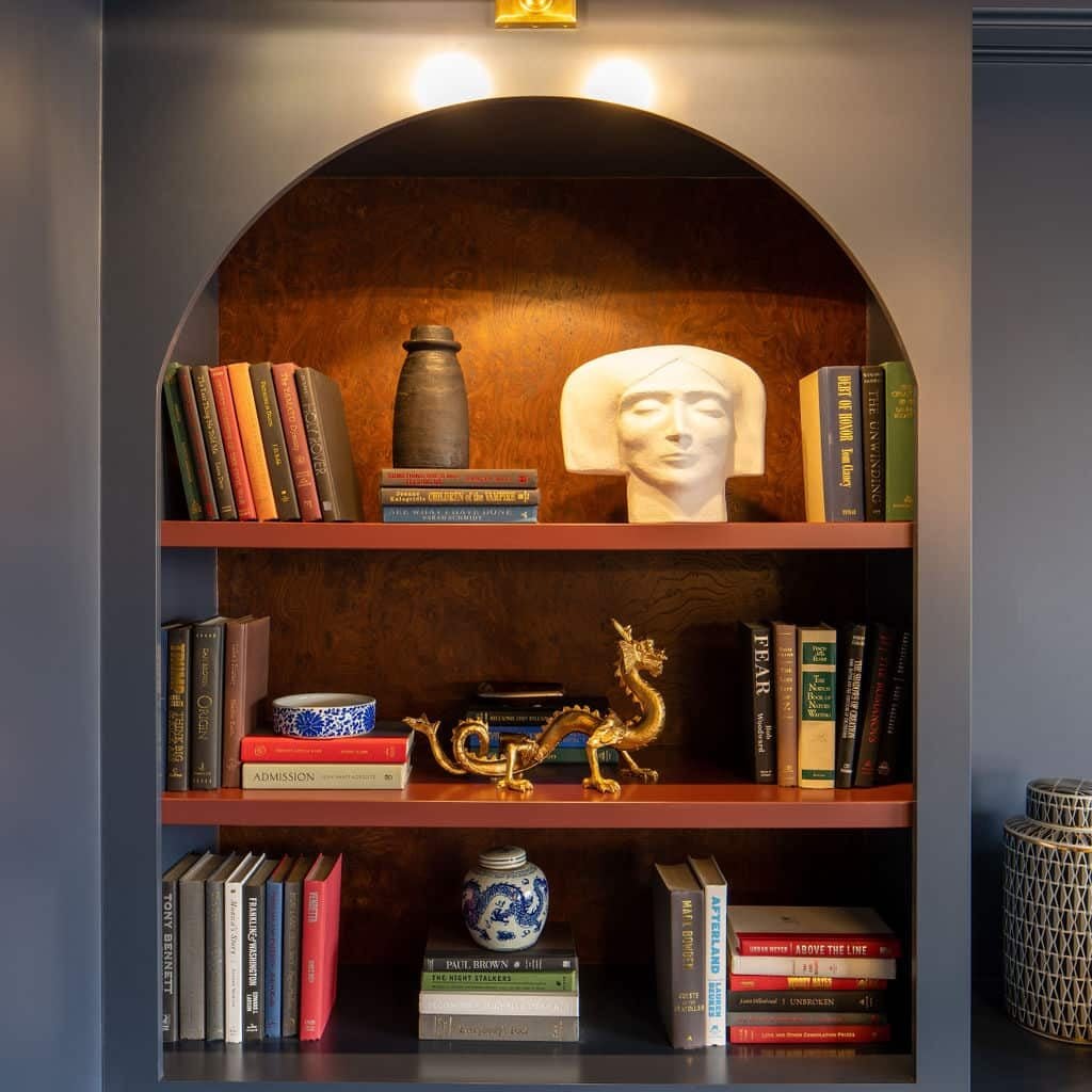 A built-in arched bookshelf by Crimson Design Group displays books, a ceramic vase, a white sculpted head, a blue and white jar, a gold dragon figurine, and a small blue dish, all illuminated by an overhead light.