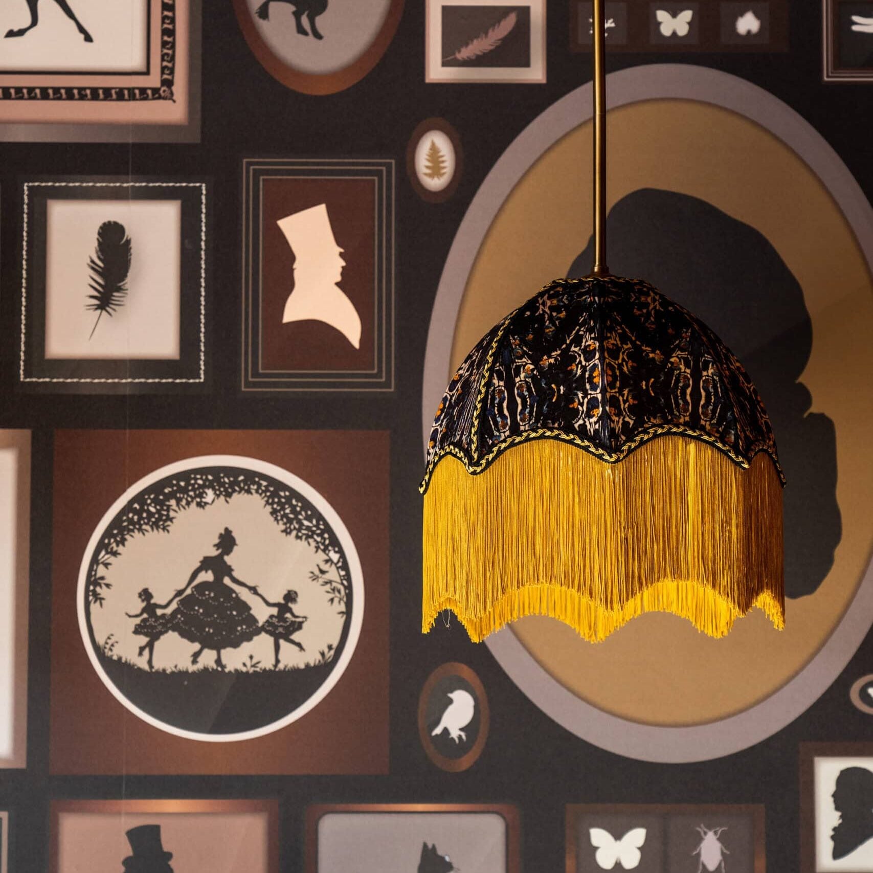 Pendant lamp with black and gold fringe hangs in front of a wall covered in silhouette artwork, including animals, people, insects, and decorative frames.