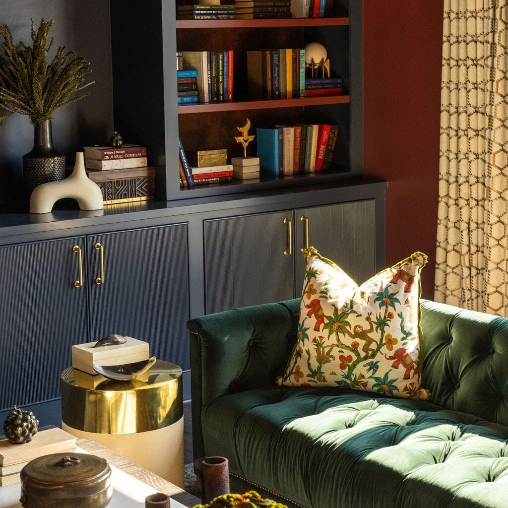 A living room with a green tufted sofa, floral pillow, gold side table, white coffee table, and built-in navy shelves filled with books, decor, and ceramics.