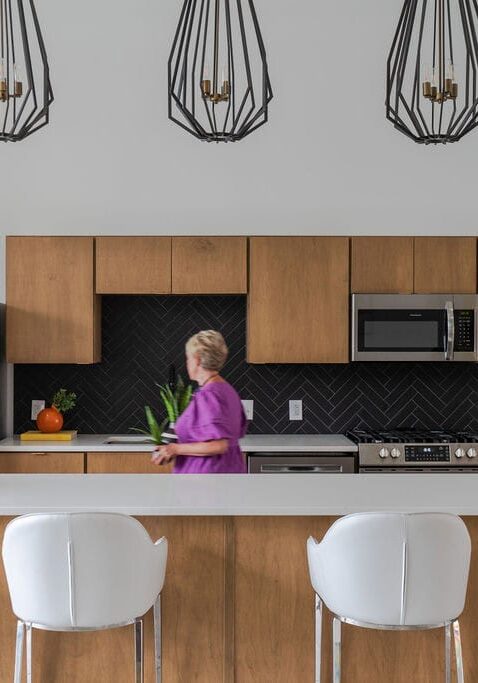 A person stands in a modern kitchen with wood cabinets, black herringbone backsplash, stainless steel appliances, and white barstools at a countertop.