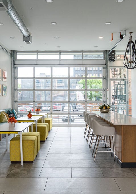 Modern communal kitchen and dining area with large windows, island seating, built-in bench, colorful pillows, and contemporary lighting fixtures.
