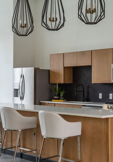 Modern kitchen with wood cabinets, stainless steel appliances, white island, four white barstools, black tile backsplash, and three geometric pendant lights.