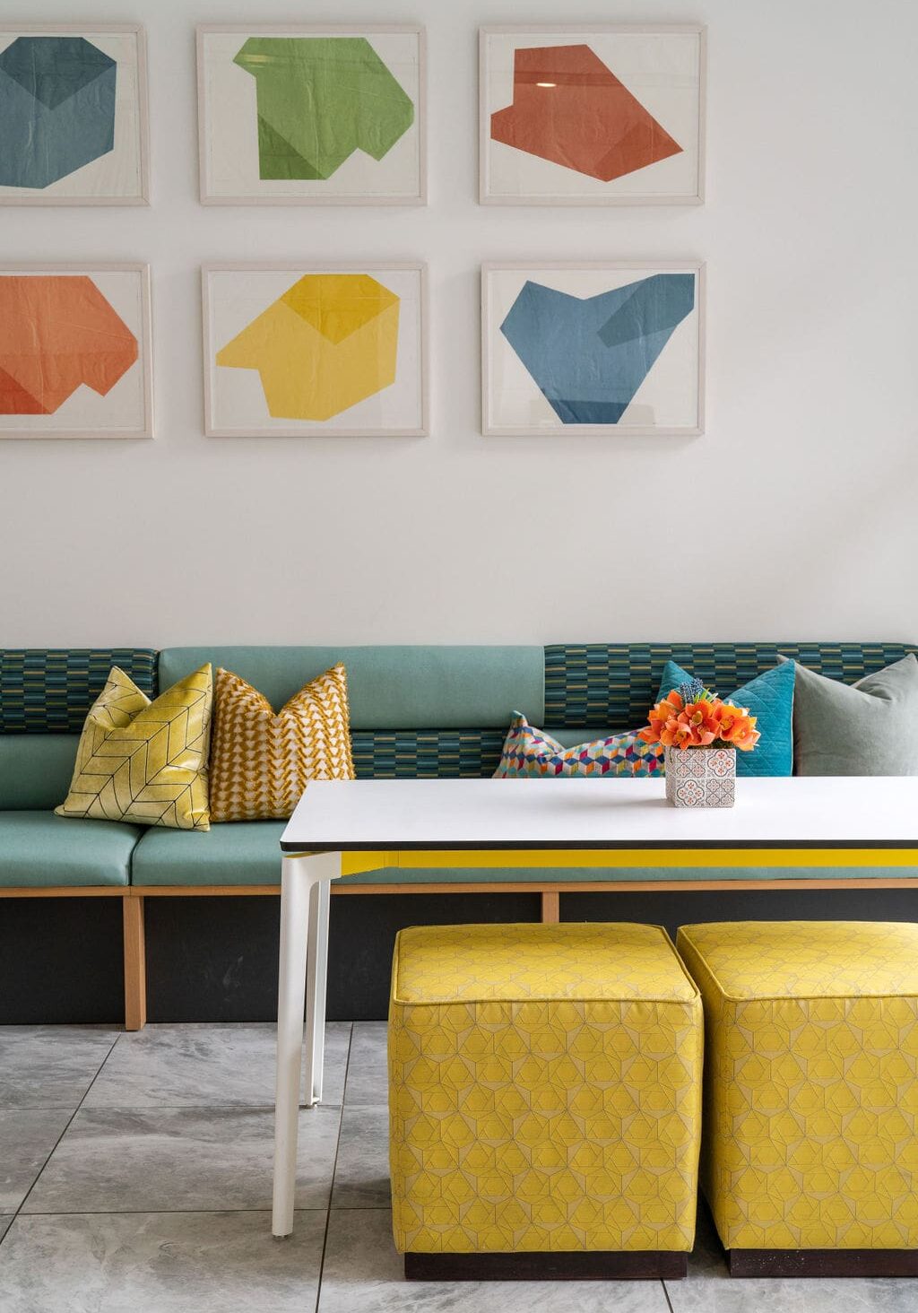 A modern seating area with a green bench, colorful pillows, a white table, yellow ottomans, a vase of orange flowers, and abstract art on the wall.