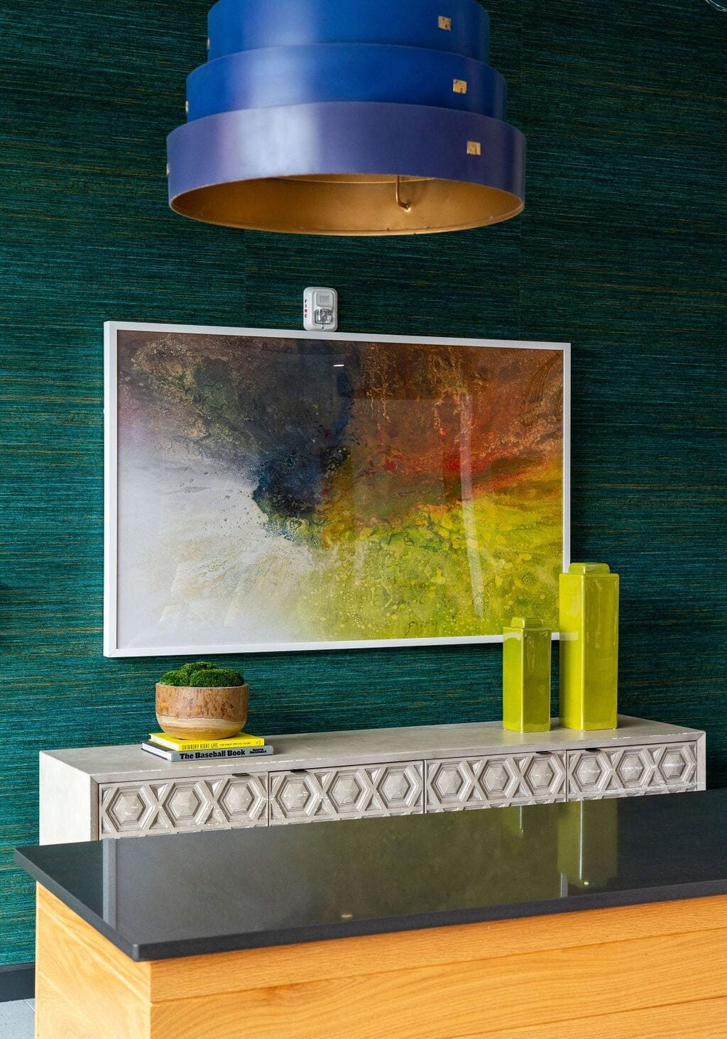A modern living space with a blue hanging lamp, abstract framed wall art, a decorative sideboard, a small potted plant, and two yellow glass vases.