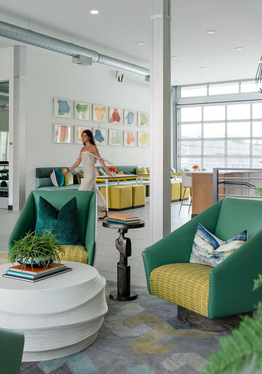 Modern lounge area with green and yellow chairs, white round table, and wall art; a woman walks in the background near large windows.
