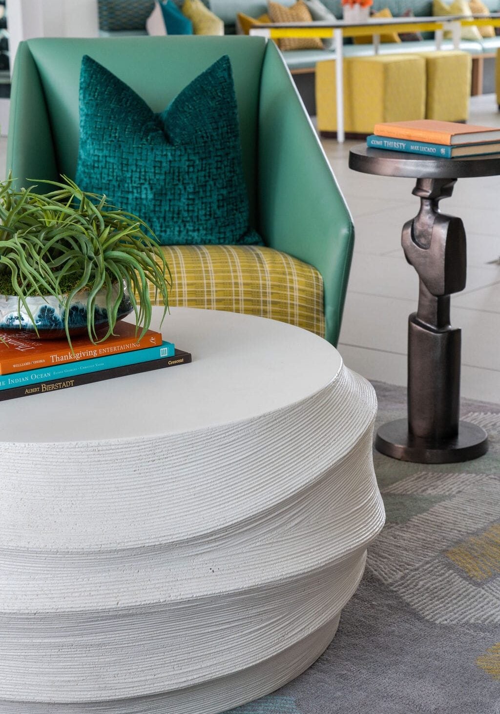 Modern living room with a round white textured coffee table, green armchair with blue pillow, small black side table, and stacked books; a potted plant sits on the table.