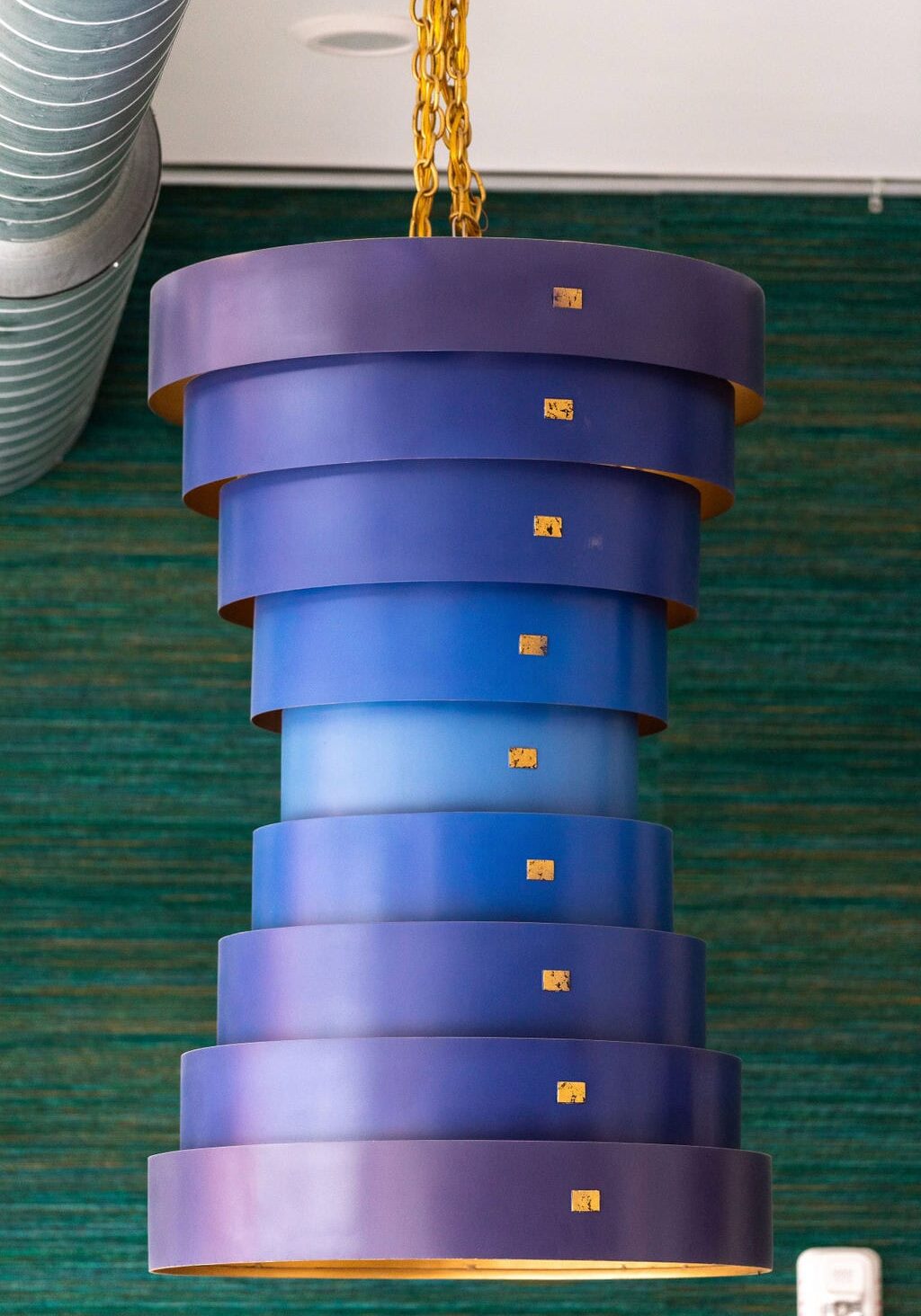 A modern, layered pendant light fixture with blue cylindrical tiers hangs from a gold chain against a green textured wall and exposed ductwork.