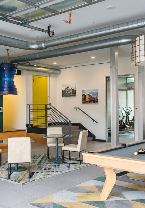 Modern lounge area with a pool table, seating, abstract art, and visible gym equipment in the background. Exposed ductwork and large windows add an industrial feel.