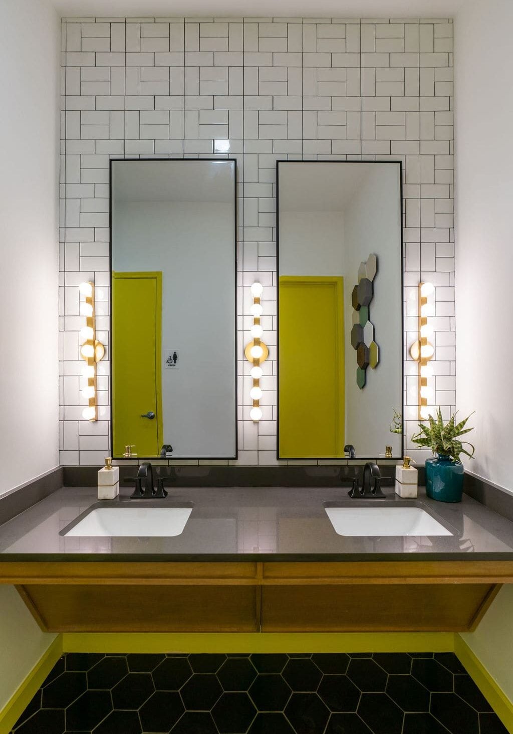 Modern bathroom with two rectangular mirrors, wall-mounted lights, white tile backsplash, double sinks, and yellow accents on doors and trim. A plant sits on the dark countertop.