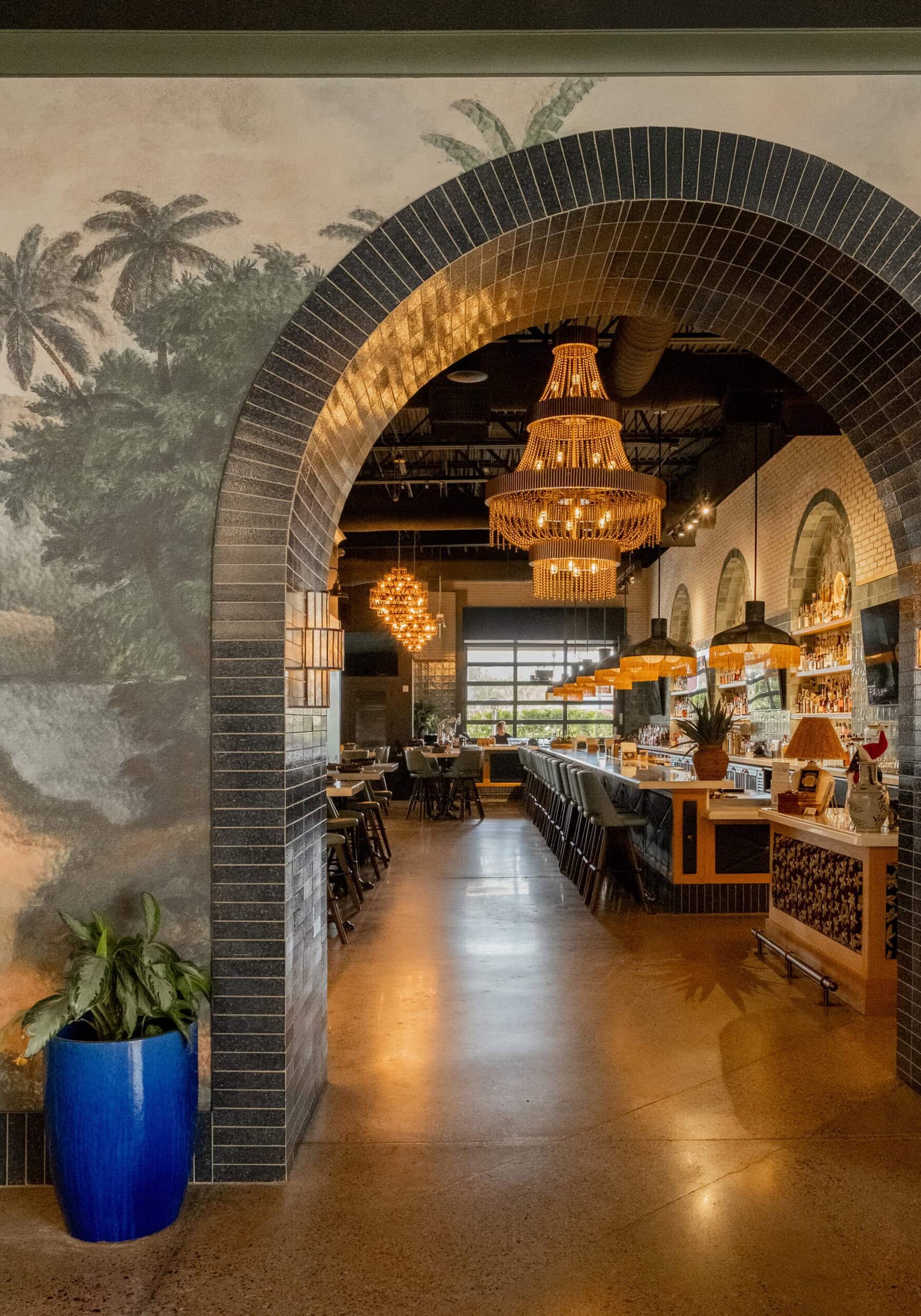 Arched entrance with black tile frames a view into a restaurant bar area, featuring chandeliers, wall art with palm trees, and a blue planter with a green plant.